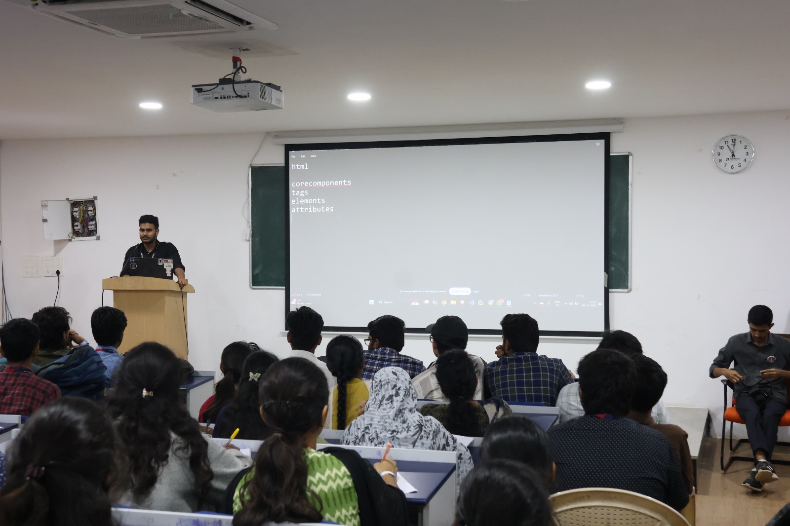 Students seated in a classroom facing a speaker and a projected list of HTML topics